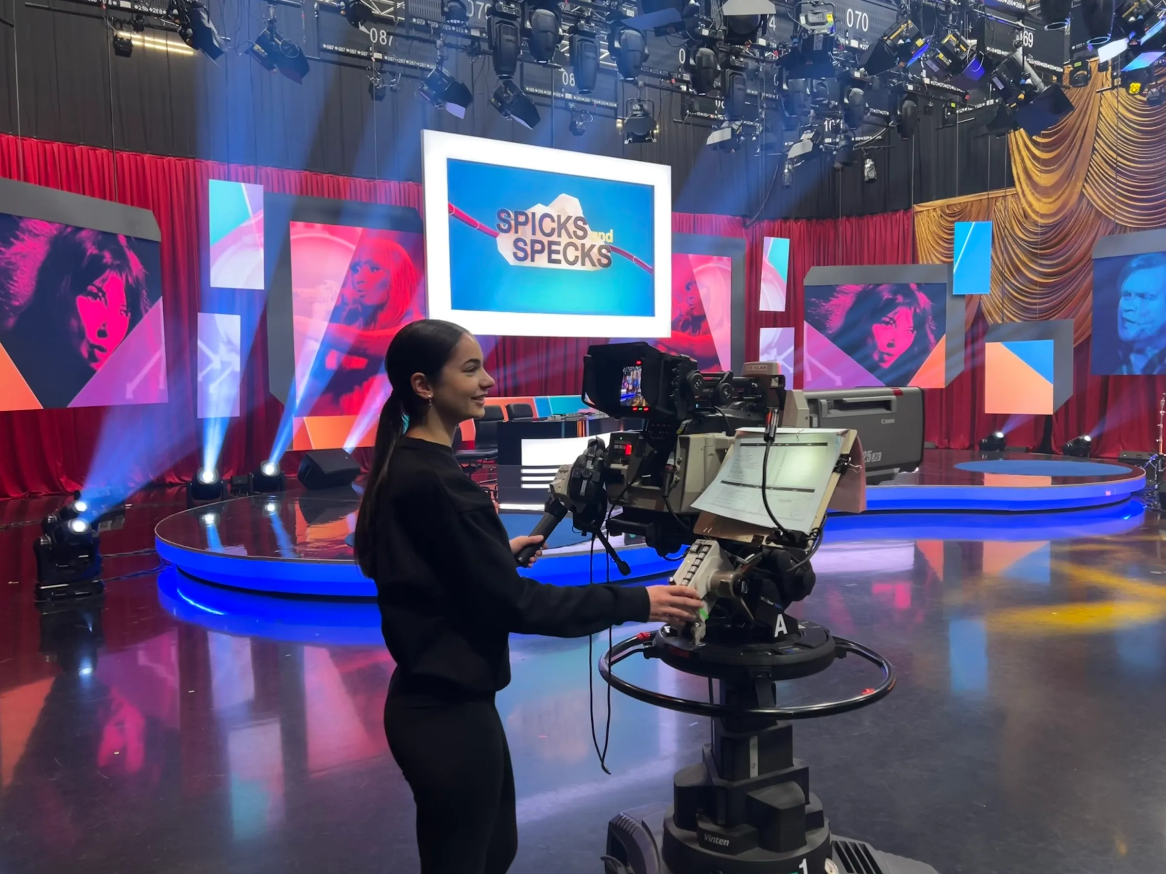Maya standing at a studio camera on the Spicks and Specks set, with colourful stage lighting and large screens behind her.