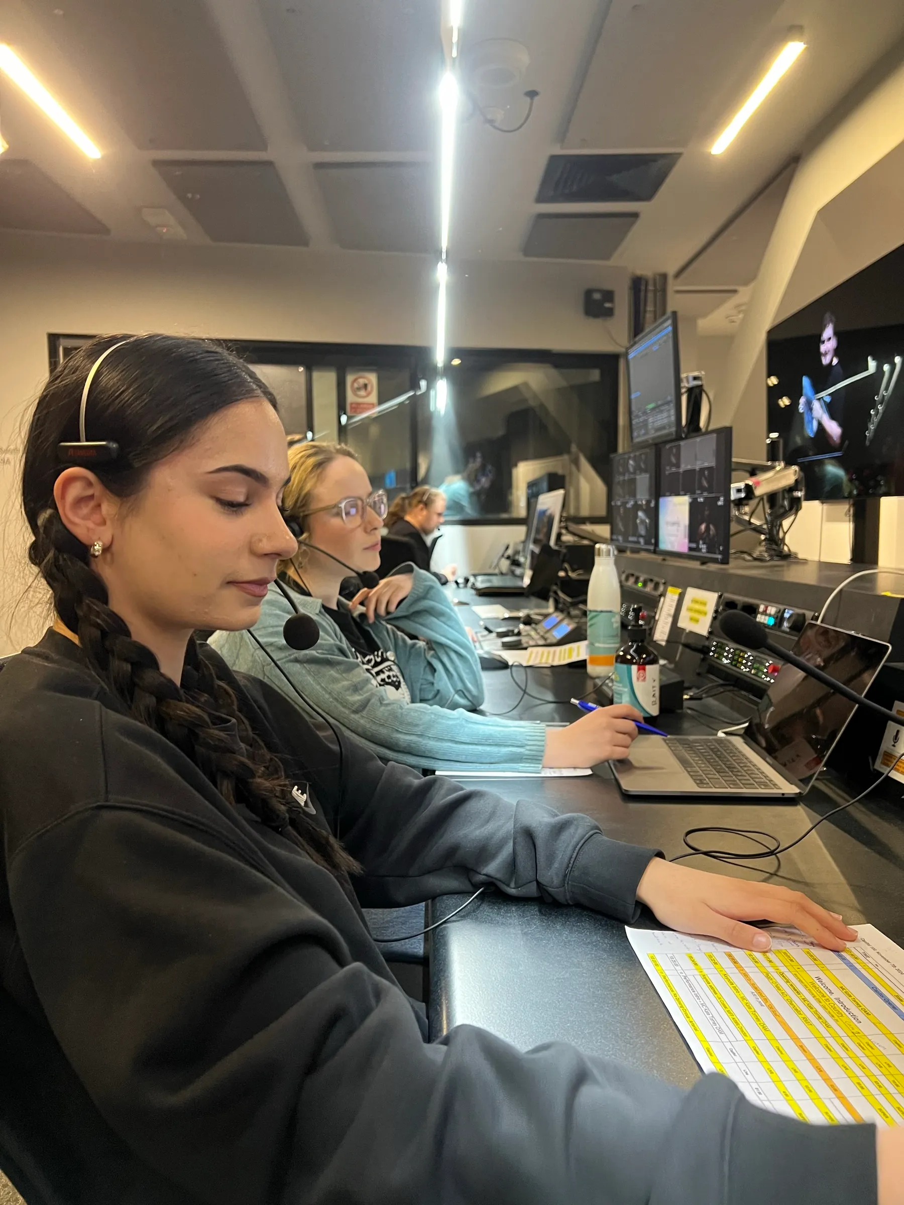 Maya wearing a headset at the control room desk during The Capitol 100 production, with a colleague beside her and a run sheet in front of her.
